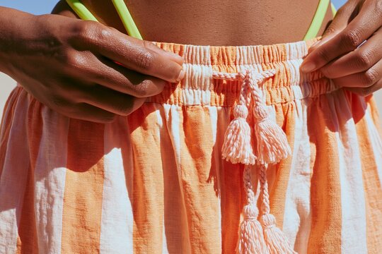 Close up of a woman's hands adjusting the waistband of an orange and white striped skirt with tassels, showcasing summer fashion and beachwear style