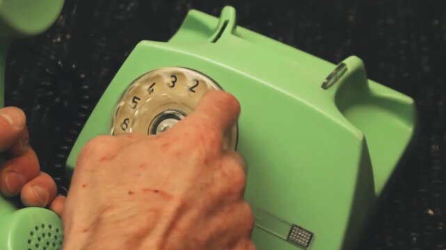 Overhead close-up hand dialing number on vintage green rotary telephone. Fingers turn dial mechanism with black numbers. Nostalgic perspective of analog communication technology from the past century