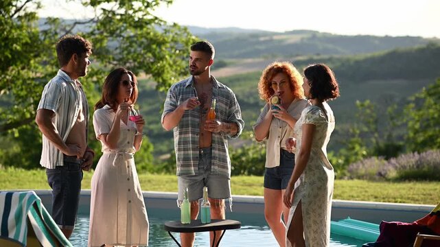 Group of young friends enjoying drinks and chatting at a summer poolside party in the countryside