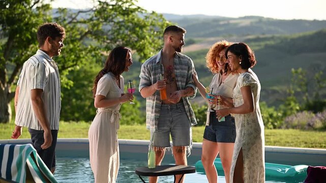 Group of friends talking and laughing together at a summer poolside party while enjoying cocktails in the backyard