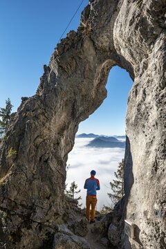 Young man stands in the rock gate Breitensteinfensterl with a view of the high fog above the valley, hiking trail to Breitenstein, Fischbachau, Bavaria, Germany