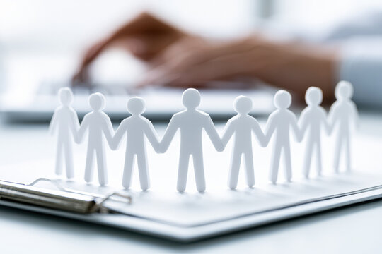 Paper cutout figures holding hands on clipboard with blurred hands typing on laptop in the background, symbolizing teamwork, unity, and collaboration.