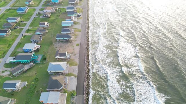 Coastal stilt frontage Surfside Beach corridor near Seashell Dr displays elevated homes bordering stone barrier beside Gulf approach. Sunlit roofs, rolling surf shimmer in Freeport, Texas