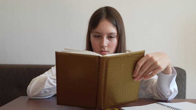 Schoolgirl thoughtfully reading book at table. Educational process and intellectual activity in study environment.