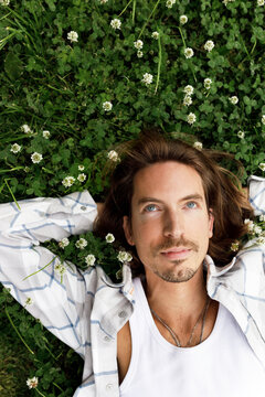 Upper vertical image of pensive thoughtful man with long hair lying on green grass with white flowers of chamomile, making wish or plans for life, being in love thinking of his crush girl
