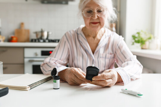 Selective focus of hands of elderly female holding glucometer checking results her blood sugar level test at home, sitting at kitchen table, suffering metabolic syndrome and insulin resistance