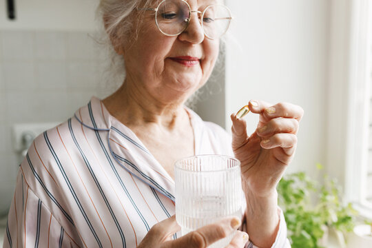 Cropped image of senior lady in glasses holding gel capsule of fish oil in one hand before swallowing as daily dietary health and antioxidant support and glass of water in another hand