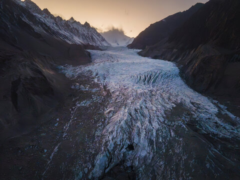 Aerial view of Pasu Glacier with its rugged ice formations and deep crevasses nestled between towering mountain peaks under a soft golden sunset light in Hunza Nagar, Gilgit, Pakistan.