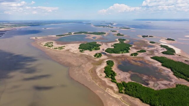 Aerial drone view of a sprawling river delta archipelago with tidal flats, sandbars, lagoons, and dense coastal wetlands beneath a vivid blue sky with scattered clouds at low tide