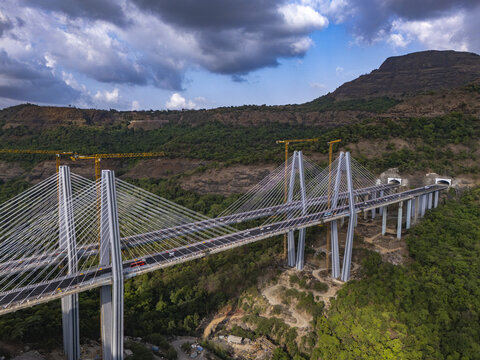 Aerial view of the Mumbai-Pune Expressway Missing Link Project featuring a cable-stayed bridge under construction with tall pylons and cranes in the hills of Khopoli, Maharashtra, India.
