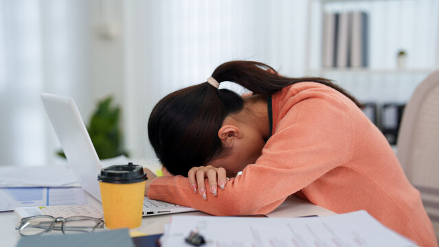 Exhausted Asian businesswoman resting head on desk beside laptop and coffee expressing work stress at home office