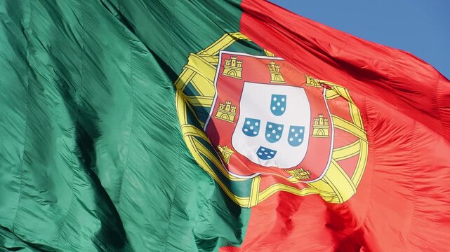 The central emblem and armillary sphere of a large Portuguese flag rippling in the wind against a blue sky background near Edward VII Park in Lisbon. Close up shot