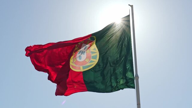 The big national flag of Portugal fluttering on a tall flagpole with the bright sun creating a strong lens flare effect from behind near Edward VII Park in Lisbon. Low angle shot