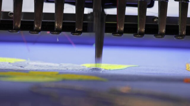Industrial embroidery machine needle stitching yellow floral pattern on blue fabric in textile factory close up.