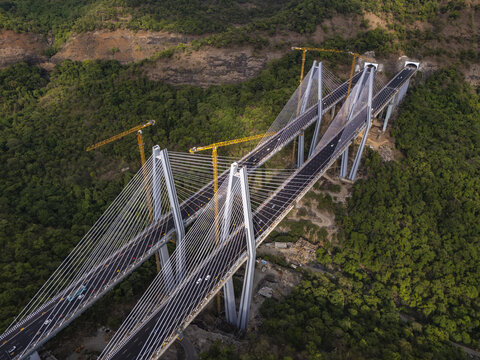 Aerial view of the Mumbai-Pune Expressway Missing Link Project featuring a cable-stayed bridge under construction with yellow cranes amidst dense forest in Khopoli, Maharashtra, India.