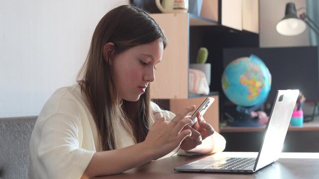 Girl sitting at table with laptop while searching for information on her smartphone.