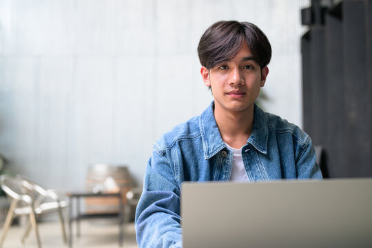 Young Asian non-binary man using laptop looking at camera wide