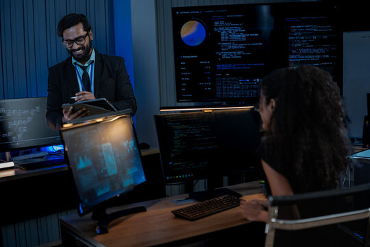 South Asian male and Black female software developers pair-program at dual monitors displaying network security algorithms at night.
