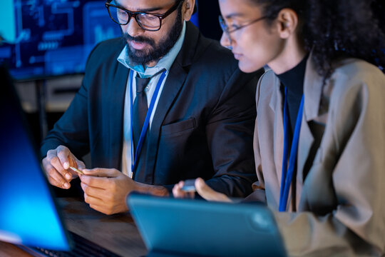 Black female data analyst cross-references code on a monitor with notes on a tablet in a blue-lit nighttime operations center.