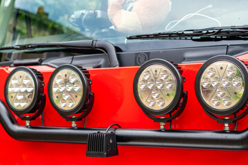 Four round auxiliary LED headlights mounted on a black bull bar on the front of a red truck or off-road vehicle, showing the windshield, wipers, and a plush toy inside. © Adam