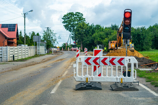 Roadworks on a suburban street, portable traffic light showing red signal, plastic white and red safety barrier, warning posts, yellow excavator working in the background, houses, cloudy sky, daylight