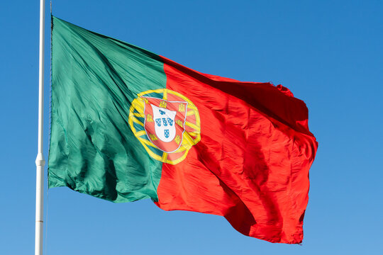 Large national flag of Portugal waving in the wind against a vibrant blue sky background. Captured from a close-up perspective near Eduardo VII Park in Lisbon