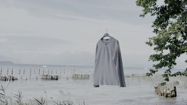 Grey long sleeve Henley shirt hanging on a hanger near a tranquil beach shoreline under a cloudy sky