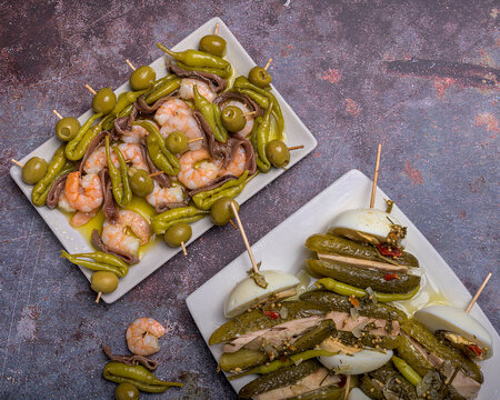 Assortment of traditional basque pintxos featuring olive and prawn gildas alongside pickle, tuna and egg skewers on plates over a dark rustic background