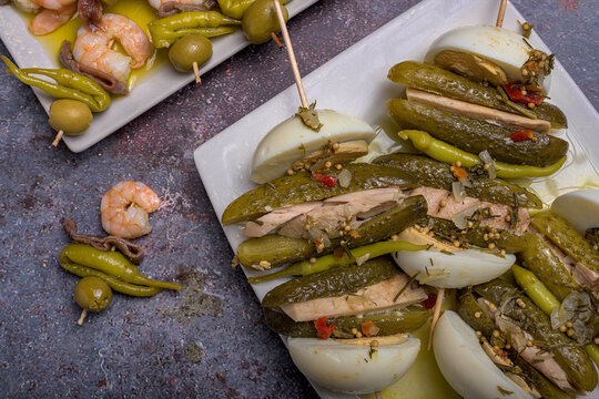 Assortment of traditional basque pintxos featuring olive and prawn gildas alongside pickle, tuna and egg skewers on plates over a dark rustic background