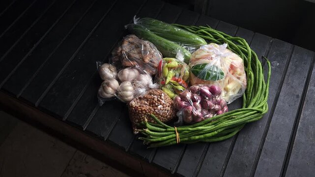 Fresh raw vegetables and cooking ingredients packaged in clear plastic bags including long beans cucumber garlic and shallots placed on a black slatted surface