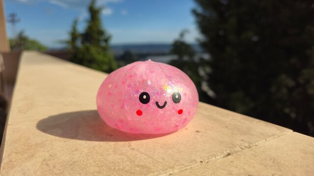 Cute pink glitter dumpling squishy toy with a smiling face sitting on a sunny stone railing outdoors.