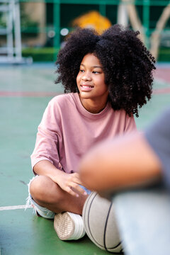Teen girl with curly hair sits on basketball court, smiling.