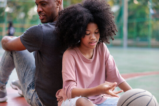 Father and daughter sit back-to-back with a basketball.