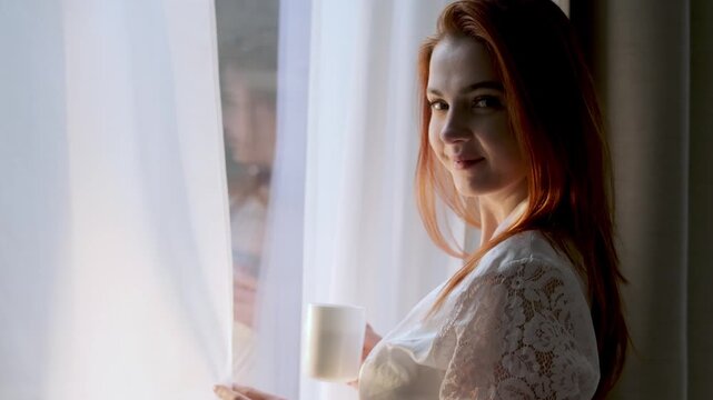 Young redhead woman in a satin robe holding a warm drink while standing by a window at home. Peaceful morning or evening moment, relaxation, mindfulness and self care lifestyle concept