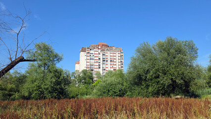 Brutalist multi story apartment block rising behind a dense green forest and a field of wild grass © hecos
