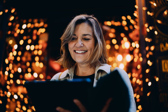 Joyful Hispanic woman using tablet outdoors at night with golden bokeh lights background, mature female freelancer smiling while looking at digital device screen, wireless communication