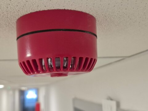 A horizontal close-up shot of a red and white fire alarm siren and strobe warning light installed on a room ceiling. Modern building safety equipment designed for fire protection and emergency evacuat