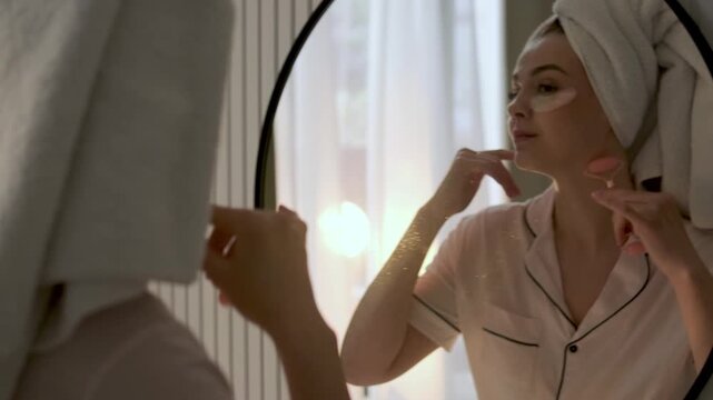 Young woman with a towel wrapped around her hair applying under eye patches while looking in a mirror at home. Morning skincare, beauty treatment and self care wellness concept