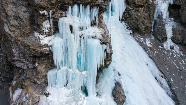 Aerial view of a frozen waterfall cascading down a rugged rocky cliff face with intricate blue ice formations and icicles in Khunjerab National Park, Gilgit, Pakistan.