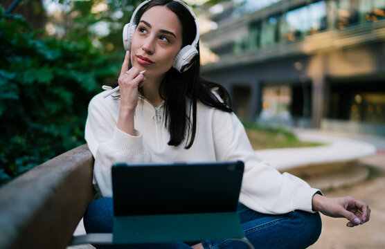 Dreamy young woman wearing headphones and holding tablet in urban park. Creative girl with braces thinking about new ideas or listening to podcast, modern digital nomad lifestyle concept.