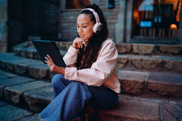 Cheerful woman with headphones uses tablet outdoors, engaged in digital activity while seated on historic steps. Combines modern tech with timeless environment. © BullRun