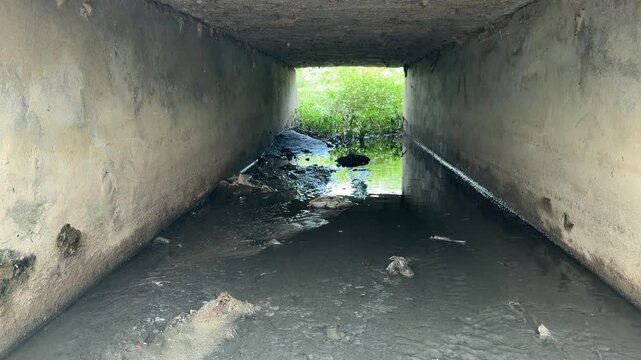 Low-angle view through dark concrete culvert tunnel with muddy stagnant water and debris, showing urban decay.