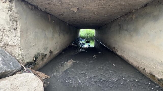 Low-angle view through dark concrete culvert tunnel with muddy stagnant water and debris, showing urban decay.