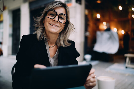 Portrait of smiling Caucasian woman in glasses using tablet at street cafe, happy middle aged professional female enjoying remote work on terrace, positive attitude and career success, bokeh.