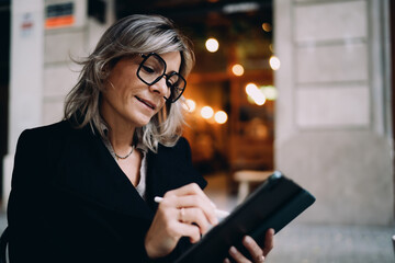 Caucasian woman in stylish glasses using digital tablet and stylus at street cafe terrace, middle aged female freelancer working remotely on portable device, connectivity and ambition. © BullRun