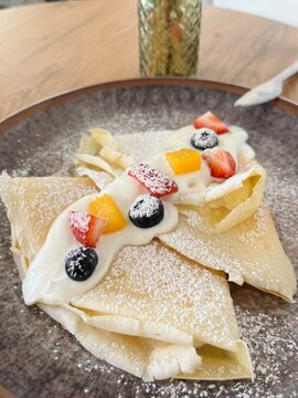 Thin pancakes folded into triangles and garnished with mixed berries, yogurt, and a light dusting of powdered sugar. Close-up food photography capturing a delicious brunch dish with colorful fruit top
