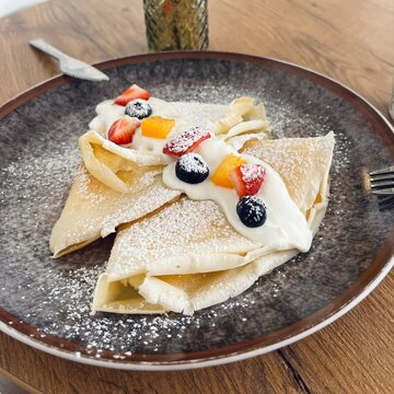Delicate folded crepes served on a ceramic plate and topped with fresh strawberries, blueberries, creamy yogurt, and powdered sugar. Elegant breakfast or dessert presentation on a rustic wooden table,