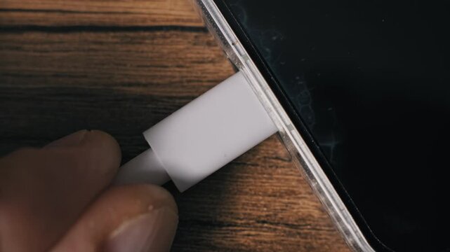 Top-down macro view of a hand inserting a white cord into a mobile phone power port. The device in a transparent shell with a dark screen rests on a textured wood grain background.