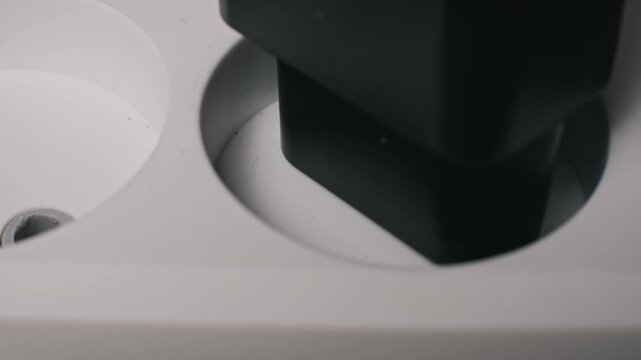 Top-down macro shot of a black Europlug with two thin prongs being inserted into a white wall socket. The electrical connection powers a household device on a wood background.