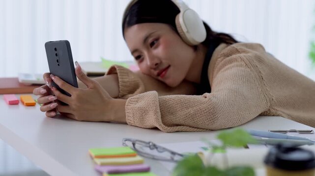 Young Asian gen z woman listening to music on headphones while browsing smartphone at modern office desk